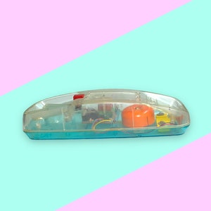 K&ouml;nnte beinhalten: Ein klares Telefon im Retro-Stil mit einem transparenten Kunststoffgeh&auml;use, das die internen Komponenten zeigt. Das Telefon hat einen t&uuml;rkisfarbenen Sockel, einen orangefarbenen Lautsprecher und eine rote Taste. Der Hintergrund ist diagonal geteilt, mit rosa und t&uuml;rkisfarbenen Abschnitten.