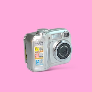 May include: Silver Nikon Coolpix 2100 digital camera with a 3x optical zoom lens. The camera has a built-in flash and a lens with the specifications 4.7-14.1mm 1:2.8-4.7. The camera is on a pink background.