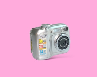 Pode incluir: Câmera digital prateada Nikon Coolpix 2100 com zoom óptico de 3x. A câmera possui flash embutido e uma lente com as especificações 4,7-14,1 mm 1:2,8-4,7. A câmera está em um fundo rosa.