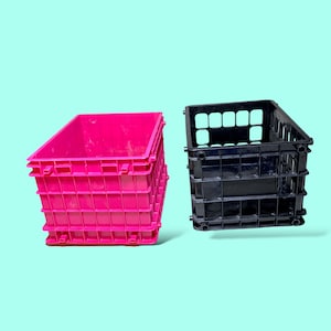Puede incluir: Dos cajas de almacenamiento de plástico. Una es rosa brillante, la otra es negra. Ambas tienen forma rectangular con un patrón de rejilla en los lados y una base sólida. Las cajas están sobre un fondo azul claro.
