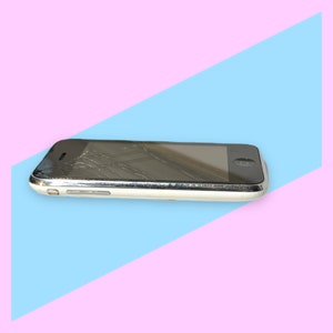 May include: A damaged, silver and white smartphone with a cracked screen. The phone is lying on a diagonal blue and pink background. The screen is black, and the phone has a silver border.