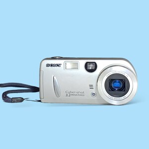 May include: Silver Sony Cybershot digital camera with a black wrist strap. The camera has a lens with a blue interior and the text "Cybershot 3.2 MEGA PIXELS". The camera is on a light blue background.