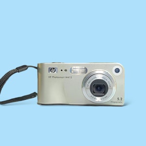 May include: Silver HP Photosmart M415 digital camera with a black wrist strap. The camera has a lens with 3x optical zoom and a 5.2-megapixel resolution. The camera is on a light blue background.