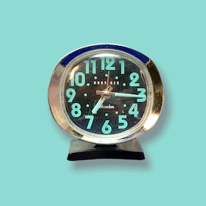 Peut inclure: Un réveil Baby Ben vintage avec un cadran noir et des chiffres et aiguilles turquoise. La montre a une lunette argentée avec un accent bleu et repose sur une base noire. Le cadran de la montre indique "Westclox".