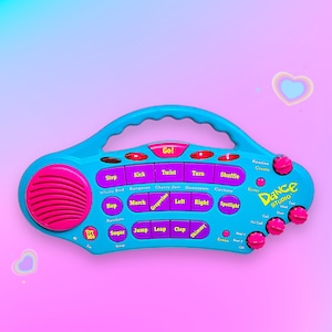Op de afbeelding: Een turquoise en roze Dance Studio-speelgoed met een handvat, knoppen en een luidspreker. De knoppen zijn gelabeld met danspassen zoals "Kick", "Twist" en "Shuffle". De tekst "Dance Studio" is afgedrukt.