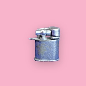 May include: A vintage, silver-colored lighter with a cylindrical body and a hinged top. The lighter shows signs of wear, with a slightly tarnished appearance. The background is a solid pink color.