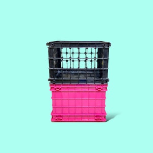 Puede incluir: Dos cajas de almacenamiento de plástico apiladas una encima de la otra. La caja inferior es de color rosa brillante y la superior es negra. Las cajas están sobre un fondo azul claro.