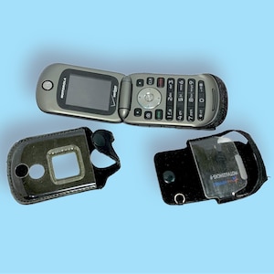 May include: A silver Motorola flip phone with a black keypad and screen, open to display the screen. Two black phone cases are also present, one with a clear window and the other with a square opening.