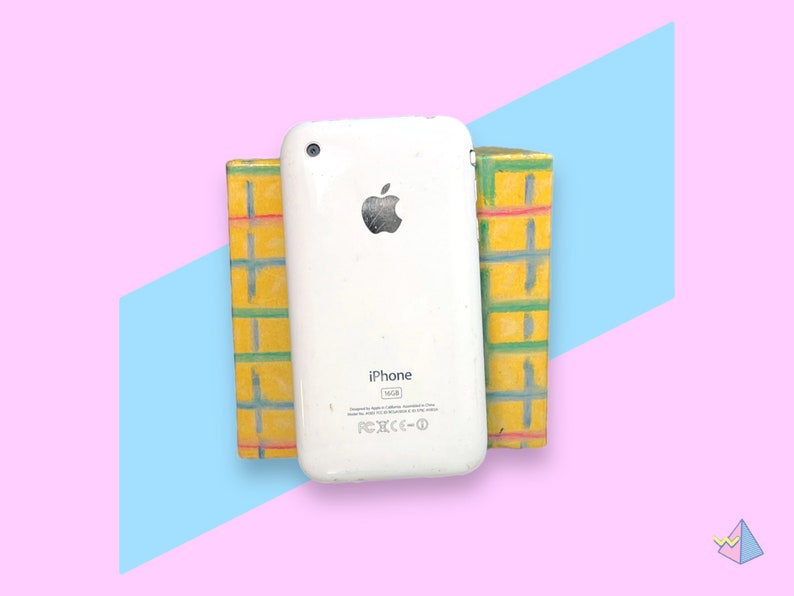 May include: A white iPhone 16GB model with the Apple logo is propped up between two yellow and green painted blocks. The background is a pastel pink with a blue and purple geometric design.