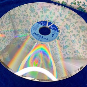 May include: A vintage DiscoVision disc with a floral pattern overlay. The disc has a reflective surface with a rainbow effect. The label reads "Blue Collar" and "1".