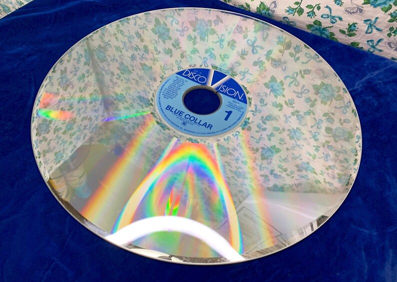 May include: A vintage DiscoVision disc with a floral pattern overlay. The disc has a reflective surface with a rainbow effect. The label reads "Blue Collar" and "1".