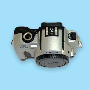 Pode incluir: C&acirc;mera Canon EOS IX Lite prateada com tampa de lente preta. A c&acirc;mera tem o logotipo da Canon e o nome do modelo impressos. A c&acirc;mera est&aacute; em um fundo azul claro.