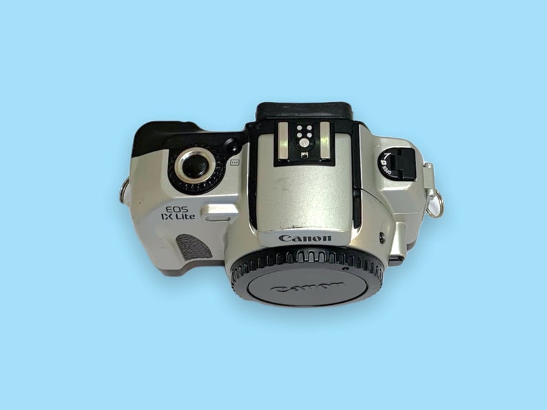 Pode incluir: C&acirc;mera Canon EOS IX Lite prateada com tampa de lente preta. A c&acirc;mera tem o logotipo da Canon e o nome do modelo impressos. A c&acirc;mera est&aacute; em um fundo azul claro.