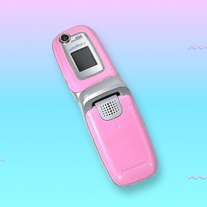 May include: Pink flip phone with a silver hinge and a small camera. The phone is open, revealing a black screen. The Sony Ericsson logo is visible on the bottom of the phone.