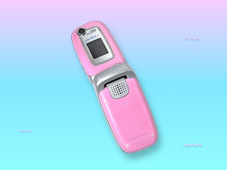 May include: Pink flip phone with a silver hinge and a small camera. The phone is open, revealing a black screen. The Sony Ericsson logo is visible on the bottom of the phone.