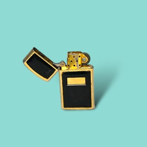 May include: A gold-colored lighter with a black rectangular panel and a hinged lid. The lighter is open, revealing the black interior. The background is a solid light blue.