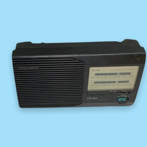 Może przedstawiać: Czarno-czarne radio Sony FM/AM z wyraźnym wyświetlaczem i pokrętłem strojenia. Radio ma prostokątny kształt z żebrowanym panelem przednim i logo Sony. Posiada przycisk zasilania i selektor pasma.