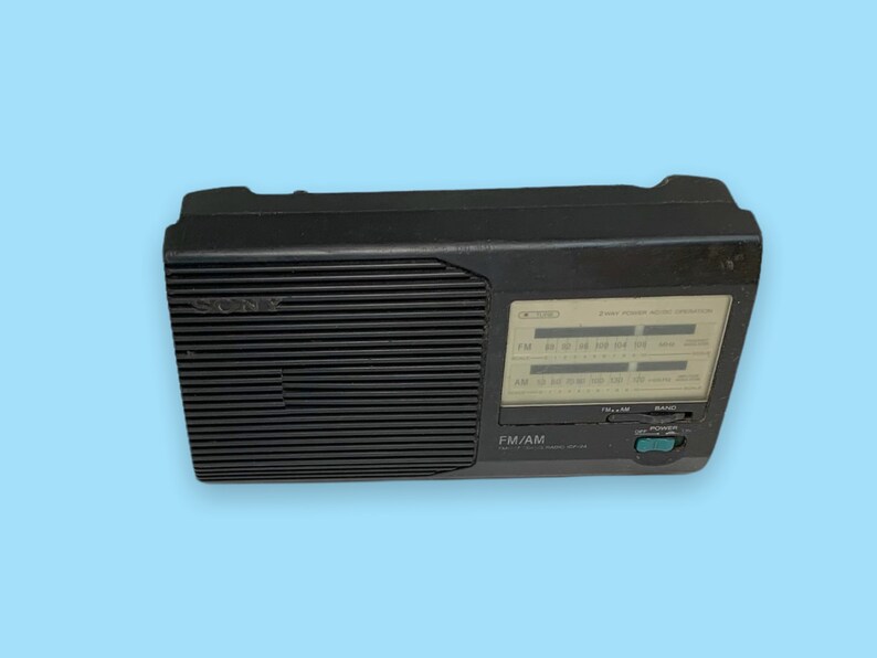 Może przedstawiać: Czarno-czarne radio Sony FM/AM z wyraźnym wyświetlaczem i pokrętłem strojenia. Radio ma prostokątny kształt z żebrowanym panelem przednim i logo Sony. Posiada przycisk zasilania i selektor pasma.