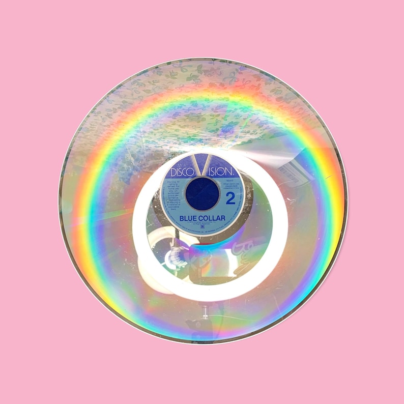 May include: A vintage Discovision disc with a rainbow effect, featuring the text "BLUE COLLAR" and the number "2". The disc is set against a pink background, creating a retro aesthetic. The image captures the iridescent quality of the disc.