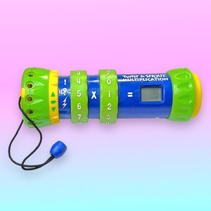 Può includere: Un giocattolo educativo blu, verde e giallo Twist & Shout Multiplication. Il giocattolo ha quadranti numerici rotanti, uno schermo LCD e un cordino con una perlina blu. Il testo "Twist & Shout Multiplication" è stampato.