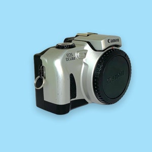 Pode incluir: C&acirc;mera Canon EOS IX Lite prateada e preta com uma tampa de lente preta. A c&acirc;mera tem o logotipo da Canon e o nome do modelo impressos. A c&acirc;mera est&aacute; em um fundo azul claro.