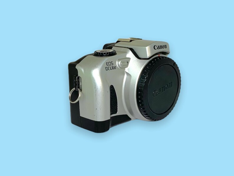 Pode incluir: C&acirc;mera Canon EOS IX Lite prateada e preta com uma tampa de lente preta. A c&acirc;mera tem o logotipo da Canon e o nome do modelo impressos. A c&acirc;mera est&aacute; em um fundo azul claro.