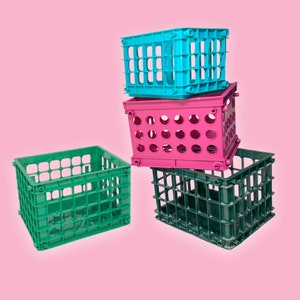 May include: Four colorful plastic milk crates stacked against a pink background. The crates are teal, pink, green, and dark green. The crates have a rectangular shape with open sides and circular or rectangular holes.
