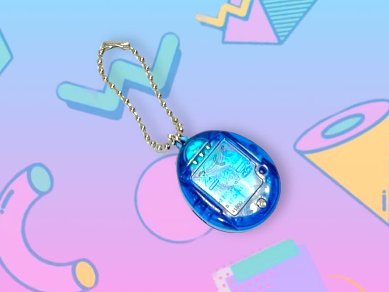 May include: A blue handheld electronic game with a silver chain. The game has a clear, oval-shaped screen with text and graphics. The background is a gradient of blue and pink with geometric shapes.