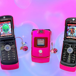 Peut inclure: Trois téléphones portables Motorola roses avec des détails noirs sont présentés sur un fond rose et violet. Deux téléphones sont ouverts, affichant des images sur leurs écrans. Chaque téléphone a un charme attaché. Les téléphones sont de style rétro.