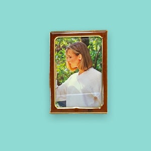 May include: A rectangular, brown picture frame with a gold trim. The frame contains a photograph of a person wearing a white sweater. The background is a light blue color.