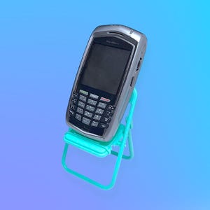 Peut inclure: Un smartphone Blackberry argenté avec un clavier complet est posé sur une petite chaise pliante turquoise. L'écran du téléphone est sombre et les touches sont noires et blanches. Le fond est un dégradé de bleu et de violet.