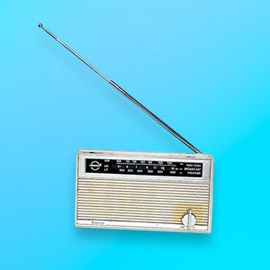 Peut inclure: Une radio vintage argentée et beige avec une antenne rétractable. La radio dispose d'un cadran avec des chiffres et un bouton de réglage. La radio est posée sur un fond bleu vif.