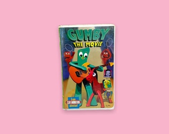 Pode incluir: Uma fita VHS vintage de "Gumby: The Movie". A capa apresenta Gumby tocando guitarra com um personagem vermelho semelhante a um cavalo. O título está em letras verdes e a fita está em um fundo rosa.