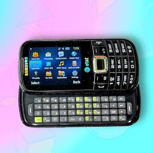 Peut inclure: Un téléphone portable Samsung noir à clapet avec un clavier coulissant, avec le logo AT&T. L'écran affiche diverses icônes d'applications. Le téléphone est sur un fond coloré. Ses dimensions sont d'environ 10 cm de long et 5 cm de large.