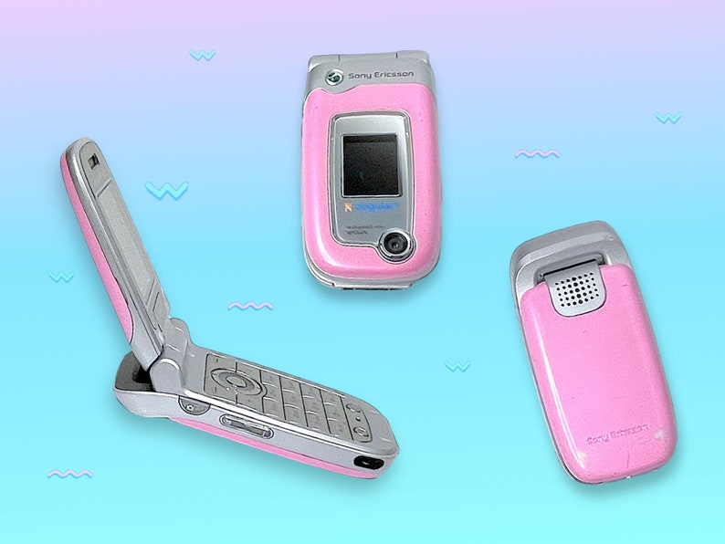 May include: Three pink Sony Ericsson flip phones are displayed against a gradient blue and purple background. One phone is open, revealing the keypad and screen. The other two phones are closed, showcasing the pink exterior and silver accents. The phones are angled to show different views.