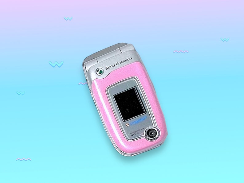 May include: A pink Sony Ericsson clamshell cell phone with a silver frame. The phone has a small screen and a camera. The background is a gradient of blue and pink with small wave shapes.