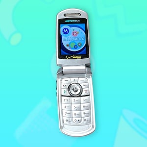 Op de afbeelding: Een zilveren Motorola klaptelefoon met het scherm open, met de tekst "HELLO MOTO" en het Verizon-logo. De telefoon heeft een toetsenbord met cijfers en functietoetsen. De achtergrond is lichtblauw.