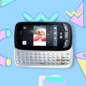 Peut inclure: Un téléphone portable LG noir avec un clavier coulissant, affichant une image d'une tortue. Le téléphone a un écran fissuré et un clavier argenté. Le fond est bleu clair avec des formes géométriques.