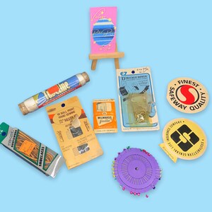 May include: A collection of vintage sewing and craft supplies, including spools of thread, shoelaces, needles, pins, and buckle rings. The items are arranged on a light blue background. Some items have text, such as "BOOT & WORK SHOELACES" and "SAFEWAY QUALITY".