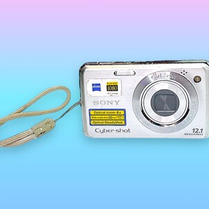 May include: Silver Sony Cyber-shot digital camera with a wrist strap. The camera features a Carl Zeiss lens, 12.1 megapixels, and optical zoom. The camera has a Full HD 1080 video setting.