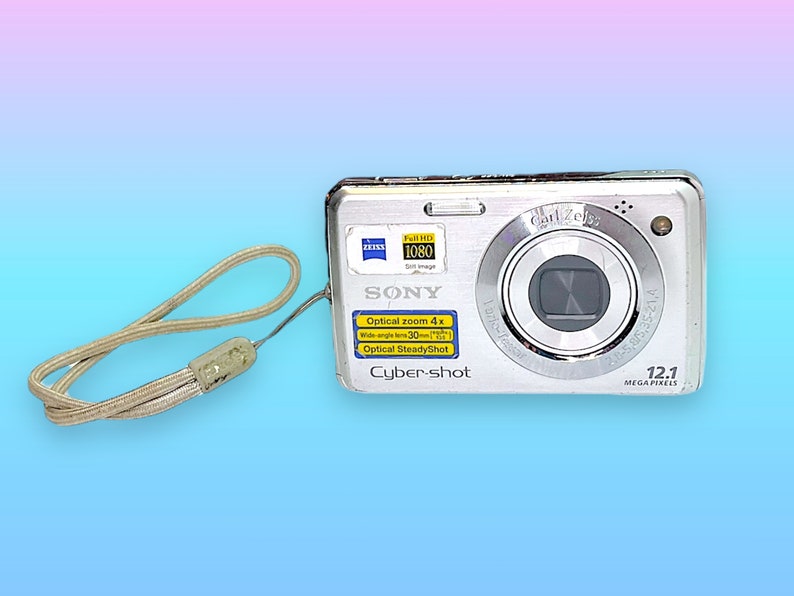May include: Silver Sony Cyber-shot digital camera with a wrist strap. The camera features a Carl Zeiss lens, 12.1 megapixels, and optical zoom. The camera has a Full HD 1080 video setting.