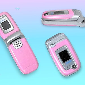 May include: Three pink Sony Ericsson flip phones are displayed against a gradient blue and purple background. The phones are open, closed, and at an angle, showcasing their silver accents, small screens, and camera lenses. The text "Sony Ericsson" is visible.