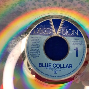 May include: A vintage MCA DiscoVision videodisc for "Blue Collar." The disc features a blue label with white text, including the title and copyright information. A rainbow reflection is visible on the disc's surface. The disc is a physical media format.