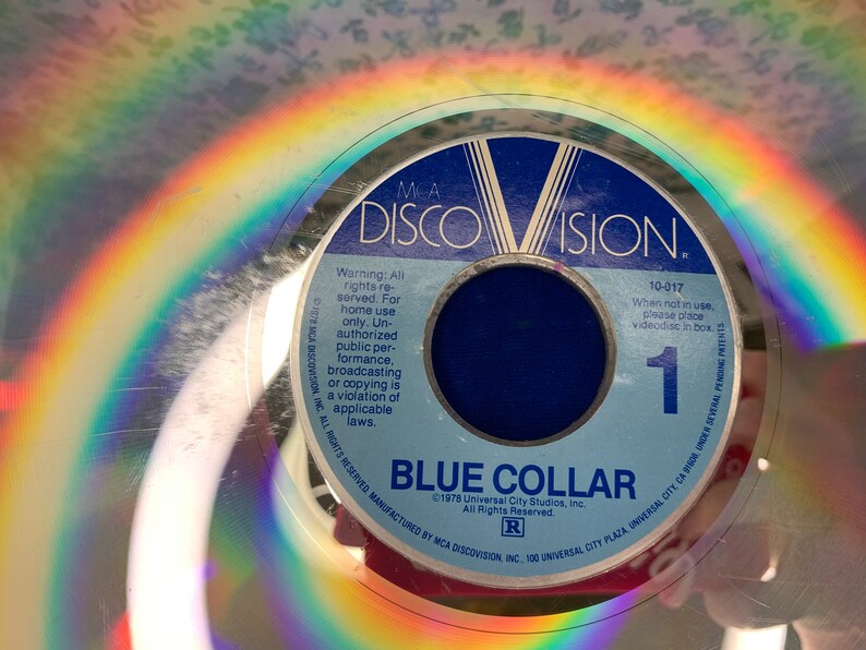 May include: A vintage MCA DiscoVision videodisc for "Blue Collar." The disc features a blue label with white text, including the title and copyright information. A rainbow reflection is visible on the disc's surface. The disc is a physical media format.