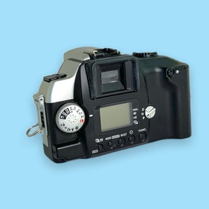 Pode incluir: Uma c&acirc;mera de filme preta e prateada com lente, visor e mostradores de controle. A c&acirc;mera tem uma pequena tela LCD e v&aacute;rios bot&otilde;es. A c&acirc;mera est&aacute; em um fundo azul claro.