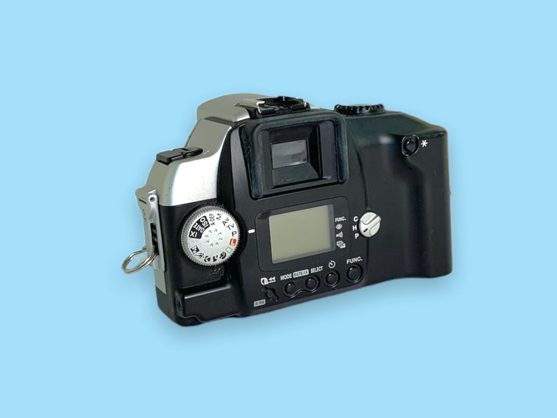 Pode incluir: Uma c&acirc;mera de filme preta e prateada com lente, visor e mostradores de controle. A c&acirc;mera tem uma pequena tela LCD e v&aacute;rios bot&otilde;es. A c&acirc;mera est&aacute; em um fundo azul claro.