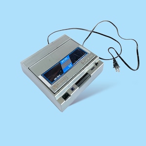 Puede incluir: Un reproductor de casetes vintage plateado y negro con un cable de alimentación negro y un enchufe de dos clavijas. El reproductor tiene una pantalla azul y negra con el texto "SOFT SHOT" y "UV-512". El dispositivo está sobre un fondo azul claro.