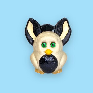 Könnte beinhalten: Ein Vintage-Furby-Spielzeug mit cremefarbenem Körper, schwarzen Akzenten und großen, ausdrucksstarken grünen Augen. Das Spielzeug hat schwarz-cremefarbene Ohren und einen gelben Schnabel. Der Hintergrund ist hellblau.
