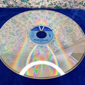 May include: A vintage Discovision disc with a floral overlay, featuring the text "BLUE COLLAR" and the Discovision logo. The disc is clear with a rainbow sheen, and the floral pattern is white with blue accents. The disc is set against a blue background.