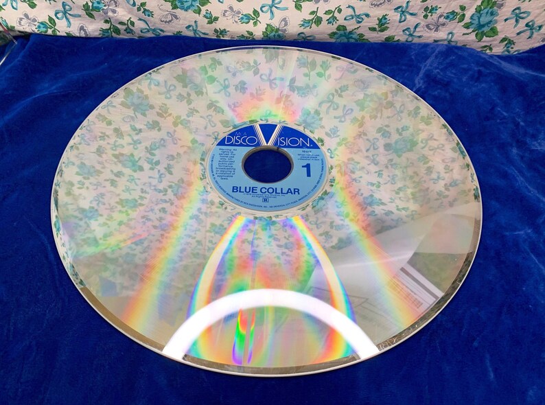 May include: A vintage Discovision disc with a floral overlay, featuring the text "BLUE COLLAR" and the Discovision logo. The disc is clear with a rainbow sheen, and the floral pattern is white with blue accents. The disc is set against a blue background.